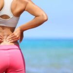 Young woman wearing athletic clothes on the beach, holding her lower back in pain