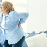 Middle aged woman in blue shirt grasping her lower back in pain.