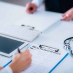 View of a desk where one set of hands is filling out health insurance paperwork across from another set of slightly blurred hands.