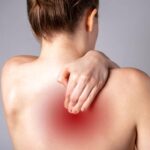 Rear view of a woman reaching over her shoulder to rub her upper back. A red spot indicates radiating pain.