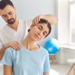 A bearded male with dark hair wearing white scrubs is manipulating the neck of a brunette woman in a light blue shirt.