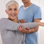Older woman stretching arm across her chest with a male in scrubs assisting her.