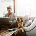 Woman sitting on couch working from home on her laptop next to her dog.