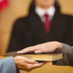 A close-up of a doctor's hands holding a pen and writing notes on a clipboard, with a blurred background of medical documents and equipment.