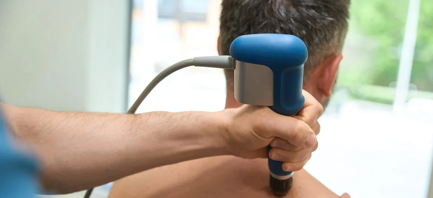 A chiropractor administers shockwave therapy to a patient's shoulder using a handheld device. The patient, seated on a treatment table, appears relaxed while the chiropractor focuses on the treatment area.