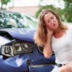 Woman standing in front of a damaged vehicle holding her neck.