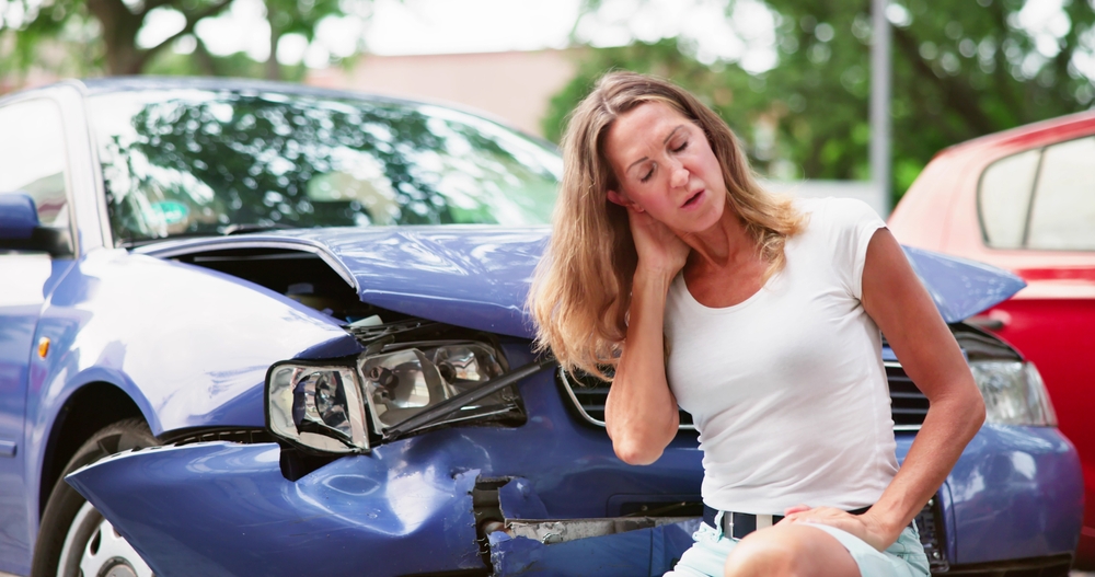 Woman standing in front of a damaged vehicle holding her neck.