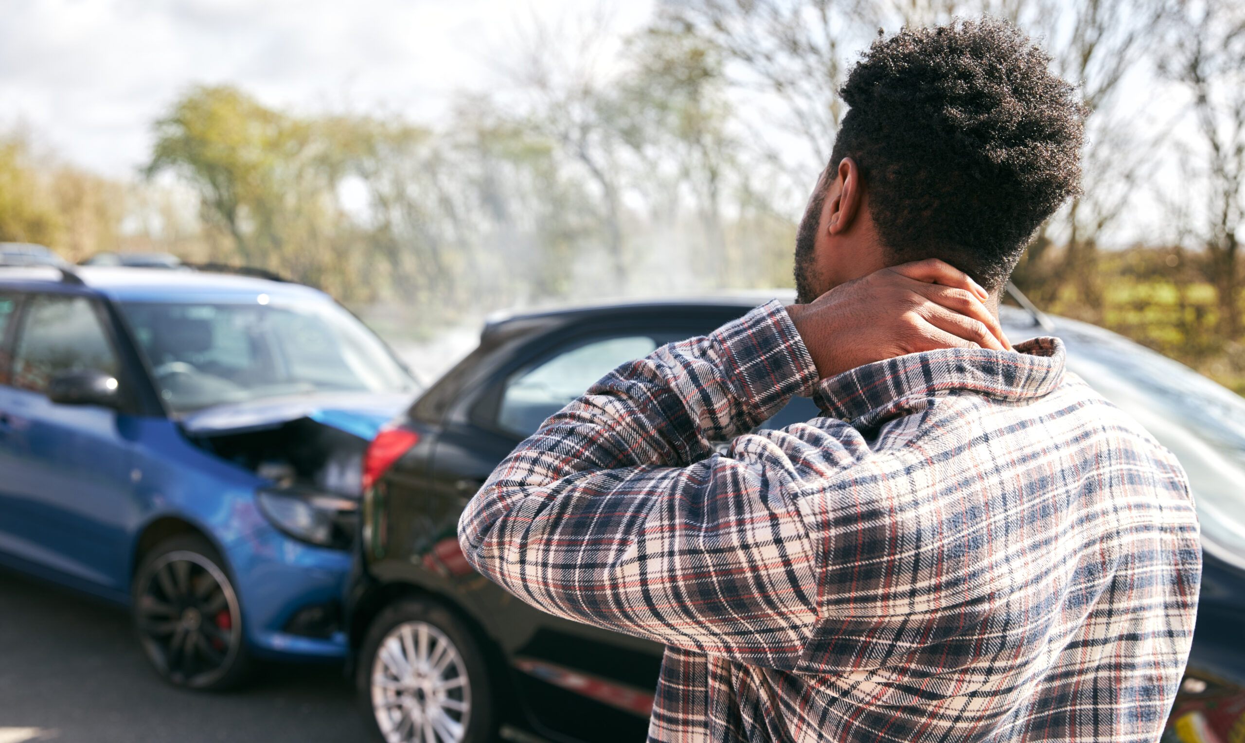 A man stands in front of two collided cars, holding the back of his neck in visible pain after an auto accident.