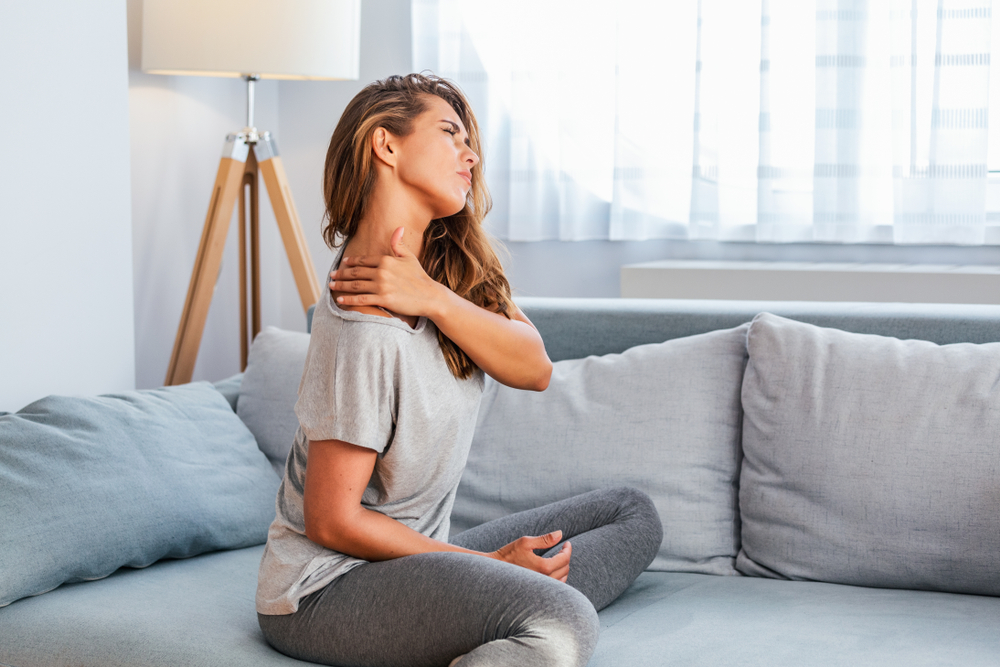 A woman sitting on a sofa holds her neck and shoulder in discomfort, seeking relief from chronic upper cervical pain in a Coral Springs clinic.