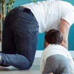 A smiling father crawling on the floor with his toddler, highlighting tips to avoid back pain for new parents.