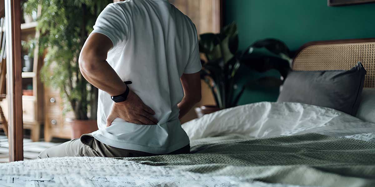 A man sitting on a bed holding his lower back, showcasing why your lower back is so stiff in the morning.