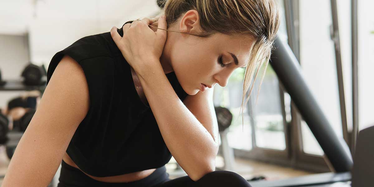 A woman in a gym holding her neck in discomfort, illustrating why your neck hurts after lifting weights incorrectly.