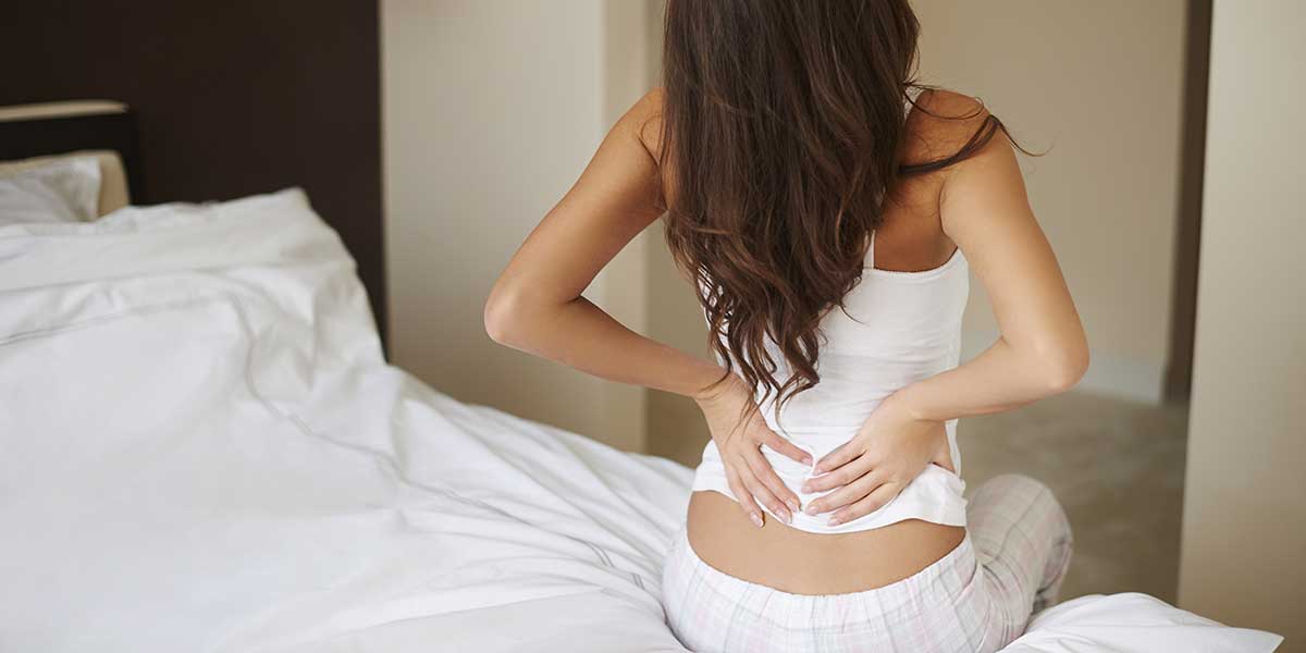 A woman sitting on her bed clutching her lower back, illustrating why sciatica feels worse in the morning hours.