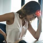 Woman slouching at office desk holding her aching head and lower back, illustrating how poor posture causes tension headaches.