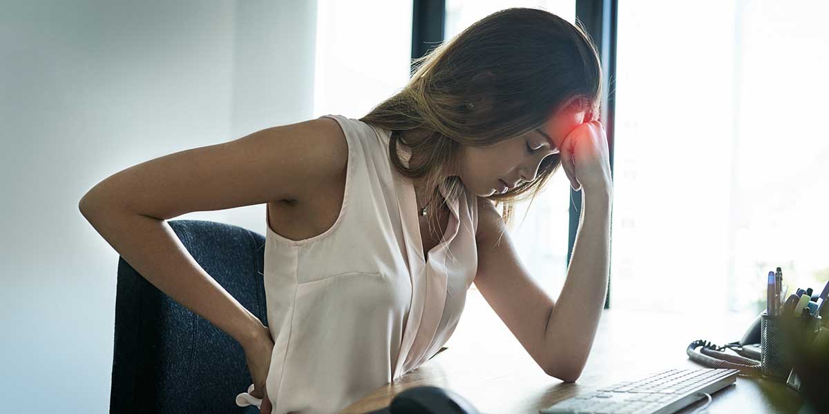 Woman slouching at office desk holding her aching head and lower back, illustrating how poor posture causes tension headaches.