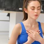 A woman sits indoors with eyes closed and hands on her chest, demonstrating deep breathing techniques to relieve back pain.
