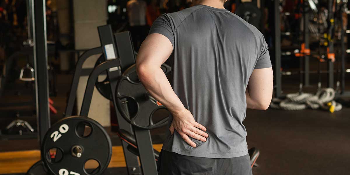 A man from behind holds his aching lower back in a gym setting near heavy weight plates after deadlifting.