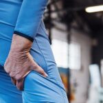 An athlete in a blue tracksuit tightly grips their painful hamstring inside a gym, highlighting sudden muscle tightness.