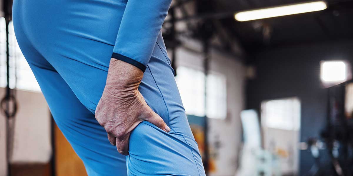 An athlete in a blue tracksuit tightly grips their painful hamstring inside a gym, highlighting sudden muscle tightness.