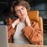 Woman sitting at an office desk clutching her painful neck, highlighting ways to prevent neck and shoulder pain from work.