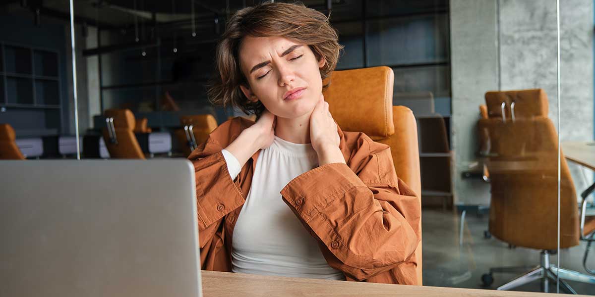 Woman sitting at an office desk clutching her painful neck, highlighting ways to prevent neck and shoulder pain from work.