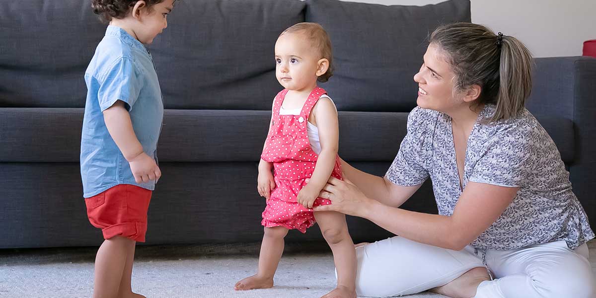 Mother helping toddler stand upright alongside another child, illustrating how good posture supports healthy physical growth in young children.