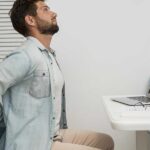 Man holding his lower back while sitting at a desk, illustrating poor posture and daily habits that trigger sciatica pain.