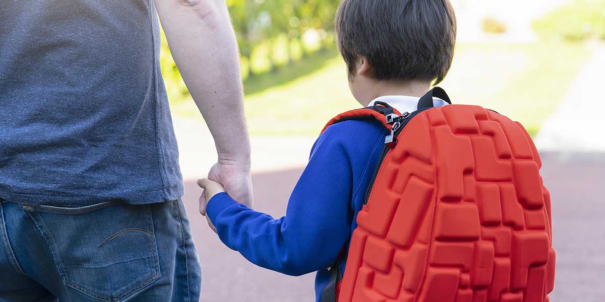 A young child wearing a bright orange backpack holds hands with an adult while walking outside toward school.