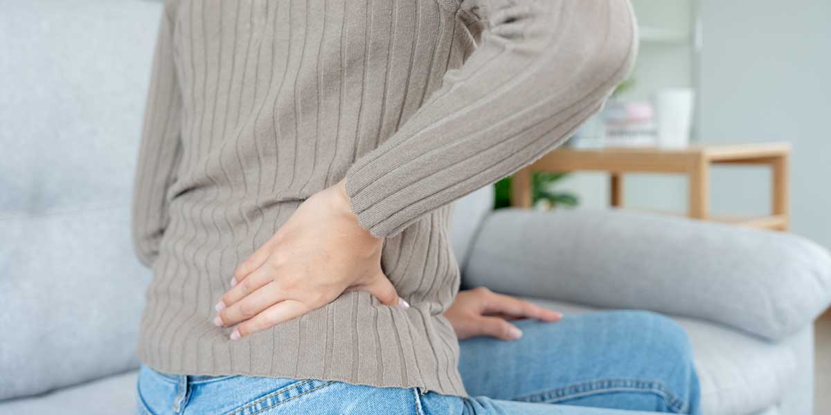 A person in a ribbed sweater holds their lower back while sitting on a couch, experiencing recurring discomfort.
