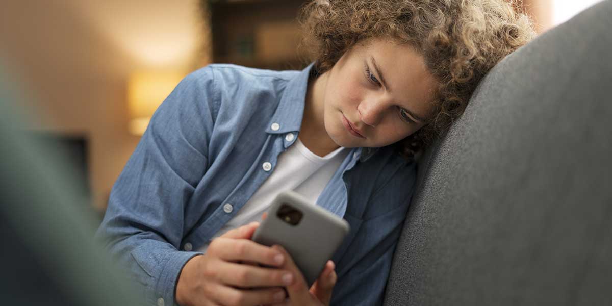 A curly-haired teenager slumps on a couch while intently looking down at a smartphone screen indoors.