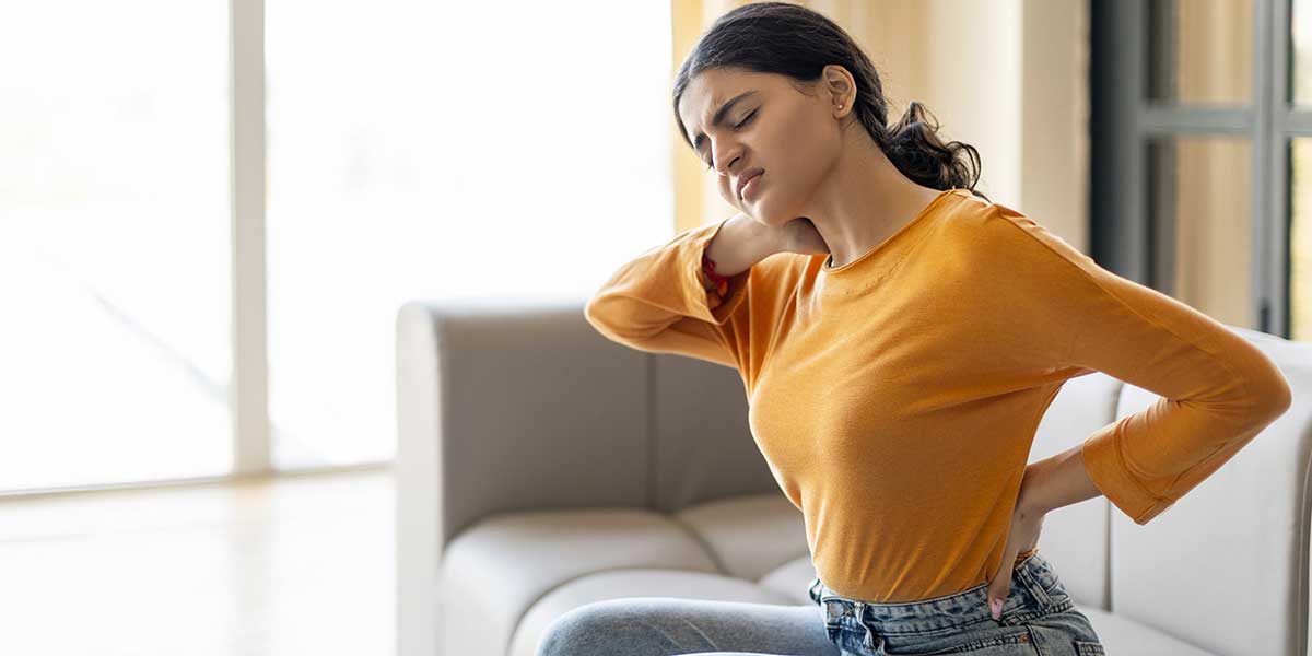 A woman wearing a yellow top holds her neck and lower back in severe pain while sitting on a couch.